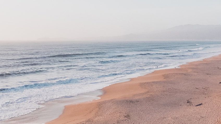 A long sandy beach with gentle waves, cliffs on the right, and distant mountains hazily visible under a soft, pale sky.
