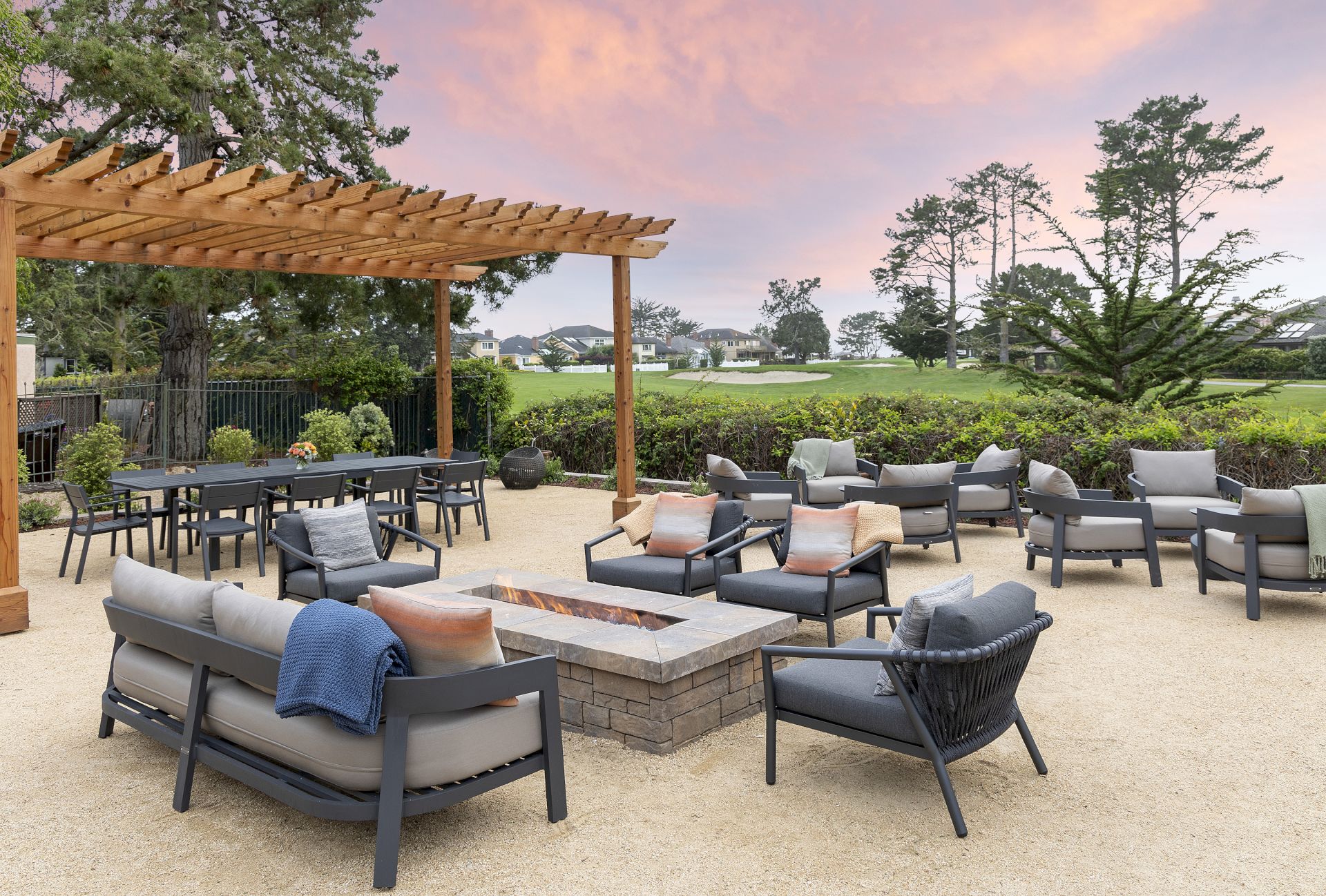 An outdoor patio with a stone fire pit, plush seating, a wooden pergola, and lush trees at sunset.