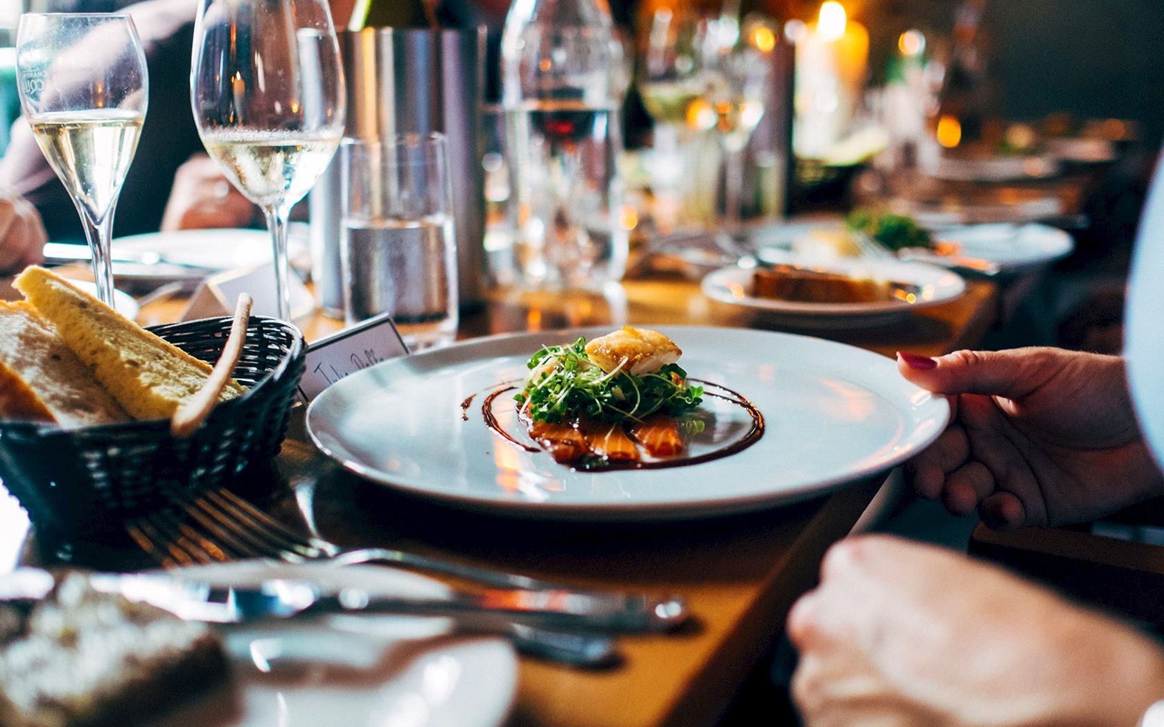 A fine dining table scene with plated appetizer, wine glasses, bread basket, and hands readying utensils for a meal.