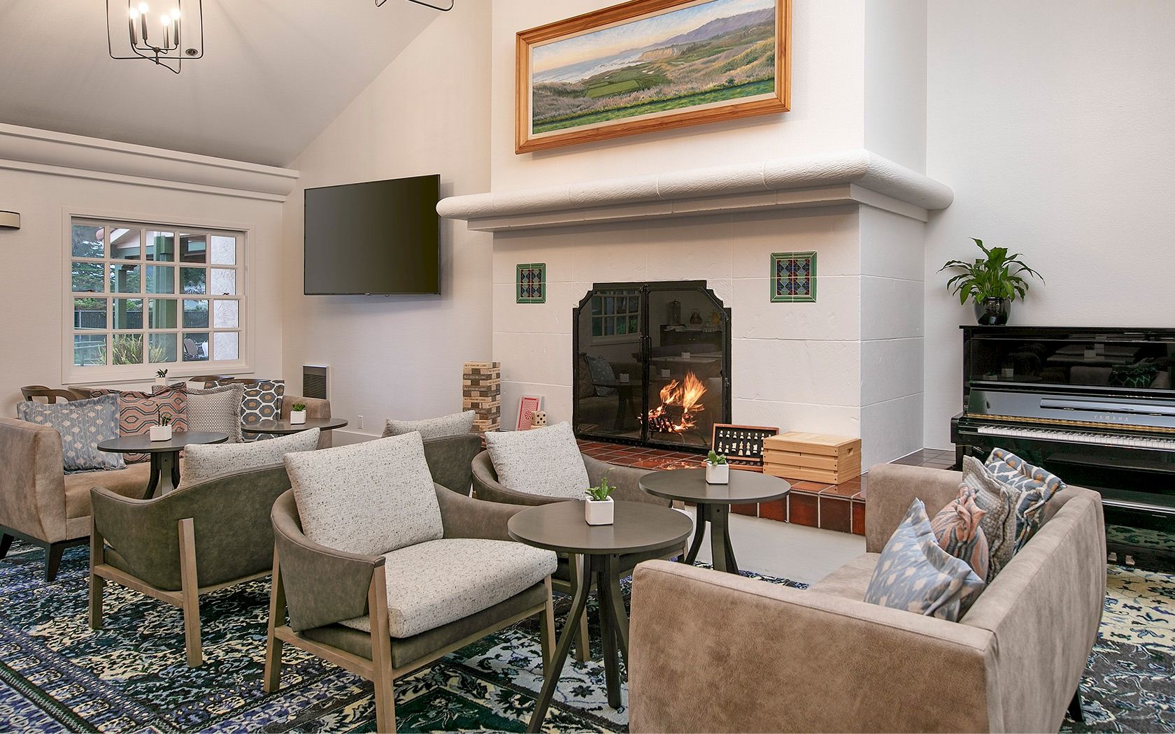 A cozy living room with a lit fireplace, glass coffee table, surrounding beige chairs, a wall-mounted TV, a piano, and a patterned rug, all in soft neutrals.