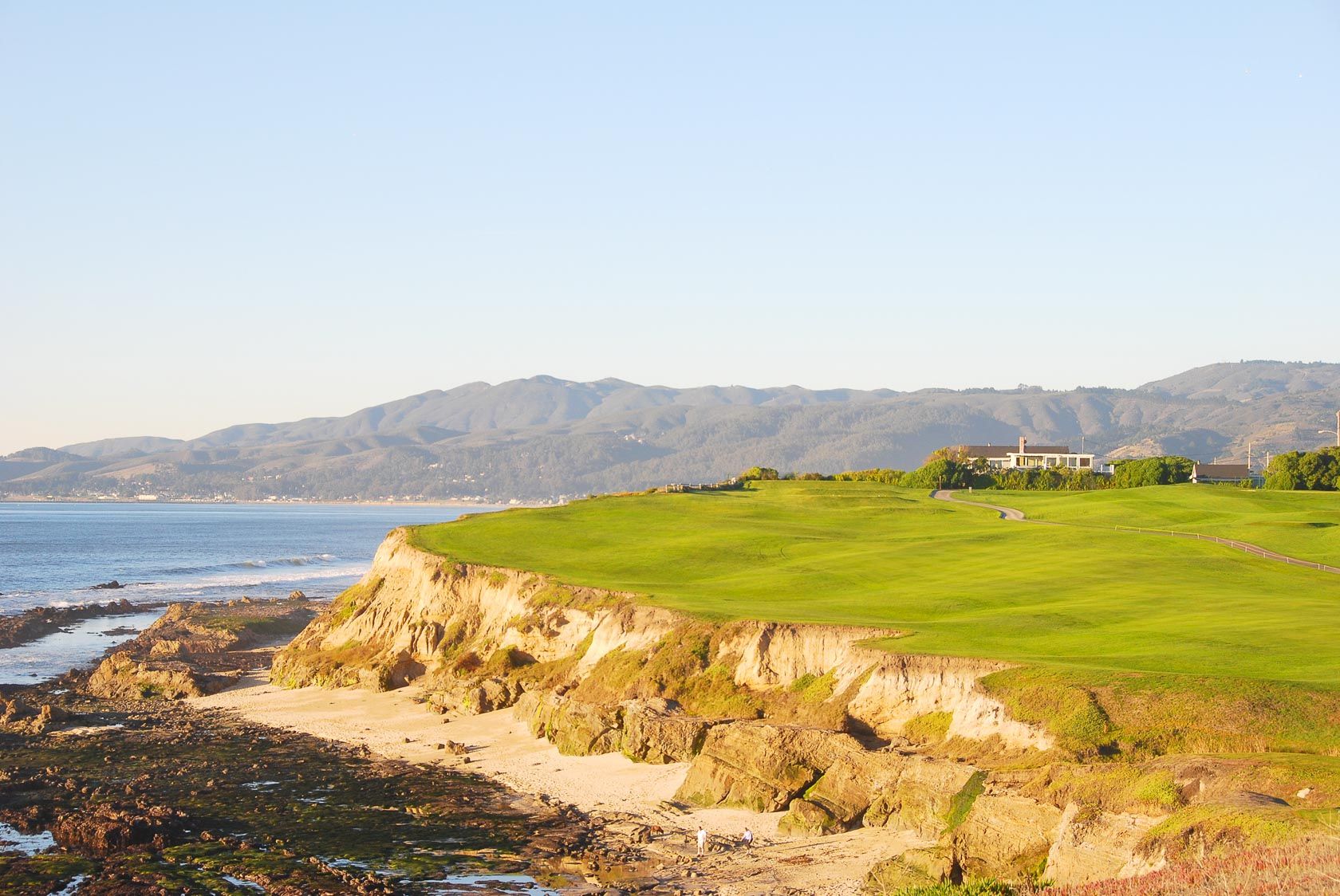 A coastal cliffside with green grassy turf, rocky shoreline, and distant hills by calm blue water, with a few buildings on the horizon.
