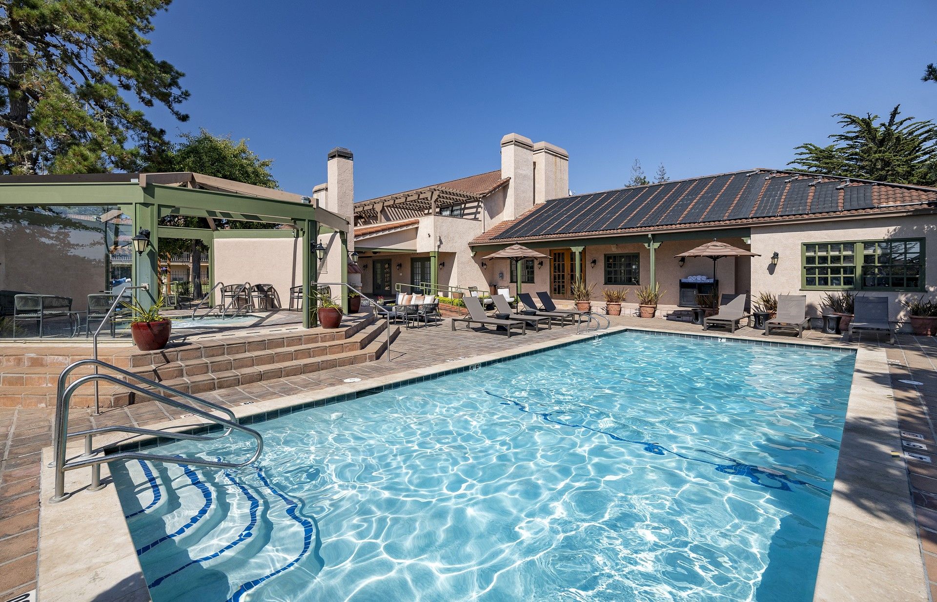 A sunny backyard with a rectangular in-ground pool, blue water, sun loungers, a patio, and a beige house with a tiled roof and solar panels, under a clear sky.
