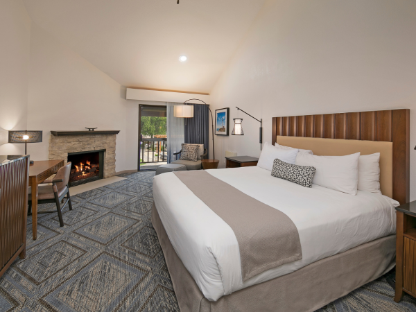 A modern hotel room with a king bed, two nightstands, a fireplace, desk, chair, and a seating area near a sliding door. Ending now.
