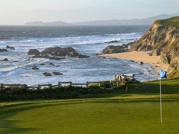 A coastal golf course with a green, flagstick by the shore, rocky cliffs, and ocean waves under a clear sky, blending sport and seascape.