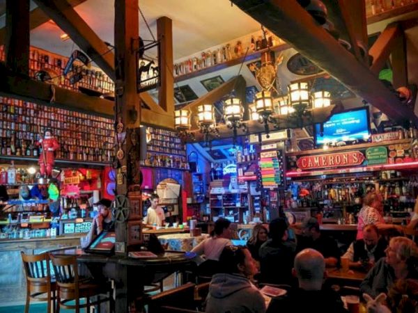 Inside a lively bar packed with colorful shelves, warm lights, a TV screen, and patrons, with a &ldquo;Cameron&rsquo;s&rdquo; sign above.