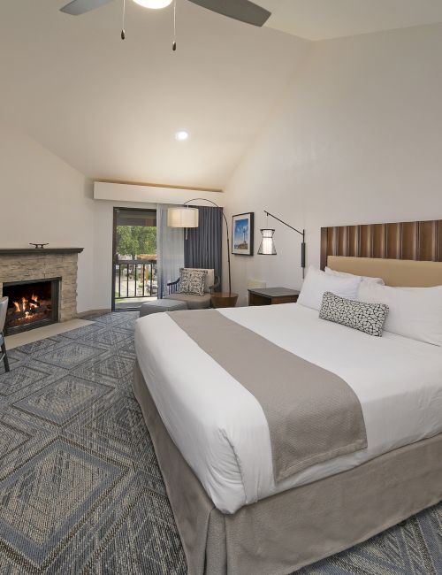 A modern hotel bedroom with a king bed, stone fireplace, TV, lamps, and large windows opening to a balcony.