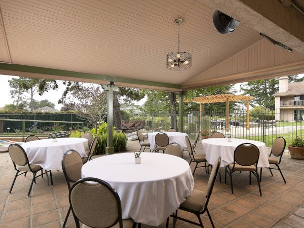 An outdoor covered patio with round tables draped in white cloths and beige chairs, set for dining in a sunny garden area.