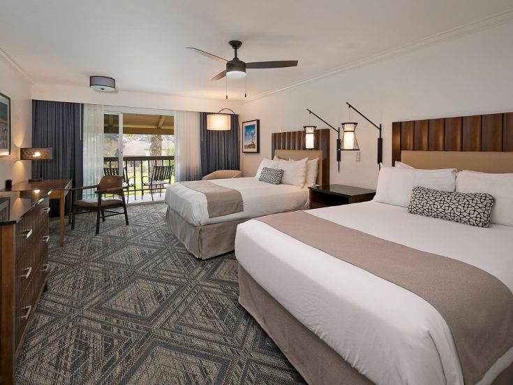 A hotel room with two queen beds, a desk, seating area, balcony, neutral decor, and ceiling fan, bright and modern.