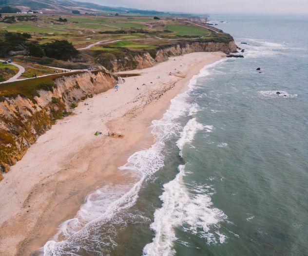 Aerial view of a sandy beach with waves, cliffs, and winding roads on a green headland under cloudy skies.