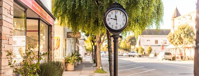 A sidewalk street scene with a decorative clock post, green trees in pots, and a storefront sign reading &ldquo;Local & Imported Wines&rdquo;.