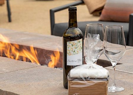An outdoor patio scene with a wine bottle, two glasses, and bread on a stone table beside a roaring fire.