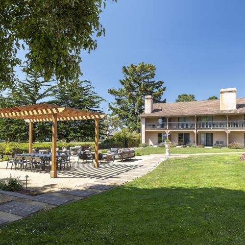 A sunny hotel-style courtyard with a green lawn, a wooden pergola over patio seating, and a two-story building in the background.
