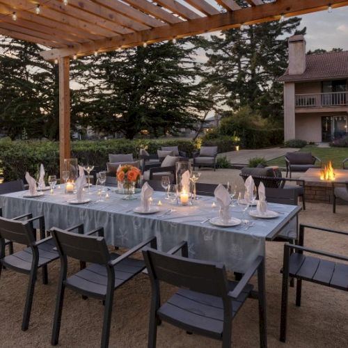 A long outdoor dining table set for a formal meal under a wooden pergola, with candles, flowers, and place settings, in a garden area.