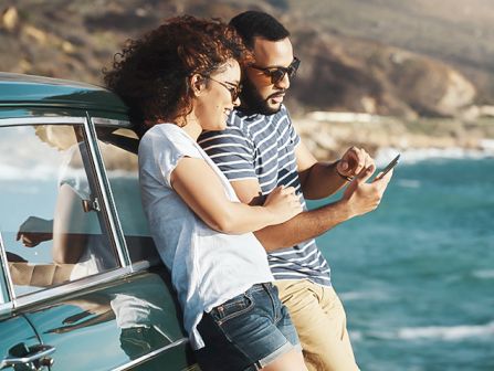 A couple leaning against a teal vintage car by the coast, looking at a tablet together as waves roll in the background.