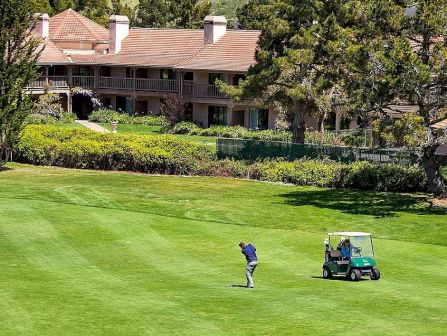 A golfer swings on a sunny course with a green cart and manicured lawns beside hillside houses.