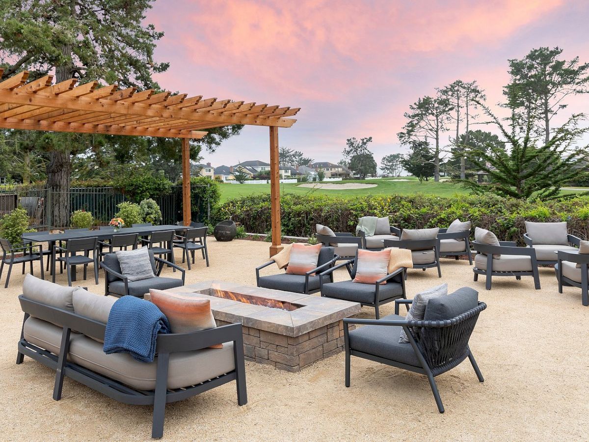 An outdoor patio lounge area with gray chairs, a stone fire pit, a wooden pergola, and a landscaped yard at sunset.