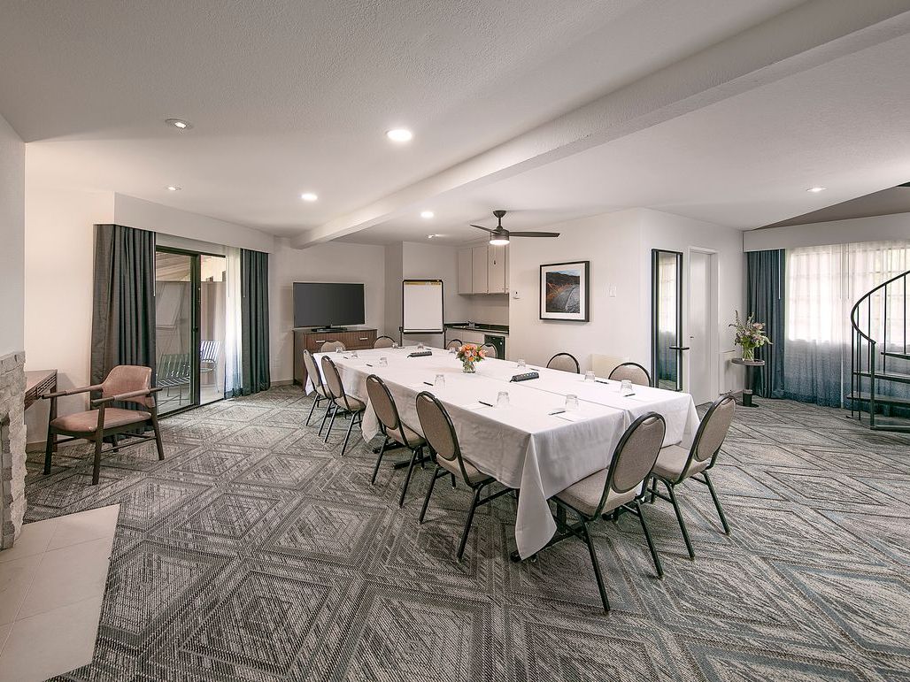 A bright conference room with a long set table, chairs, TV, kitchen pass-through, windows, and a fireplace.