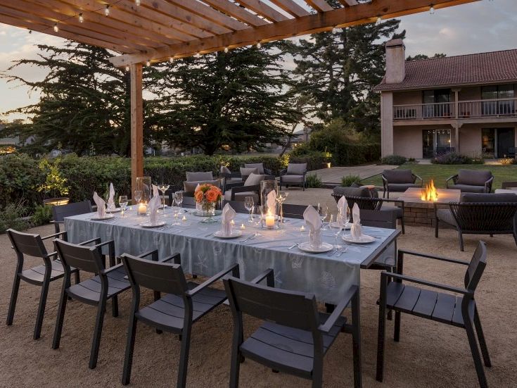 An outdoor patio banquet set: table with candles and orange flowers, cushioned chairs, and a lit fire pit beside a house.