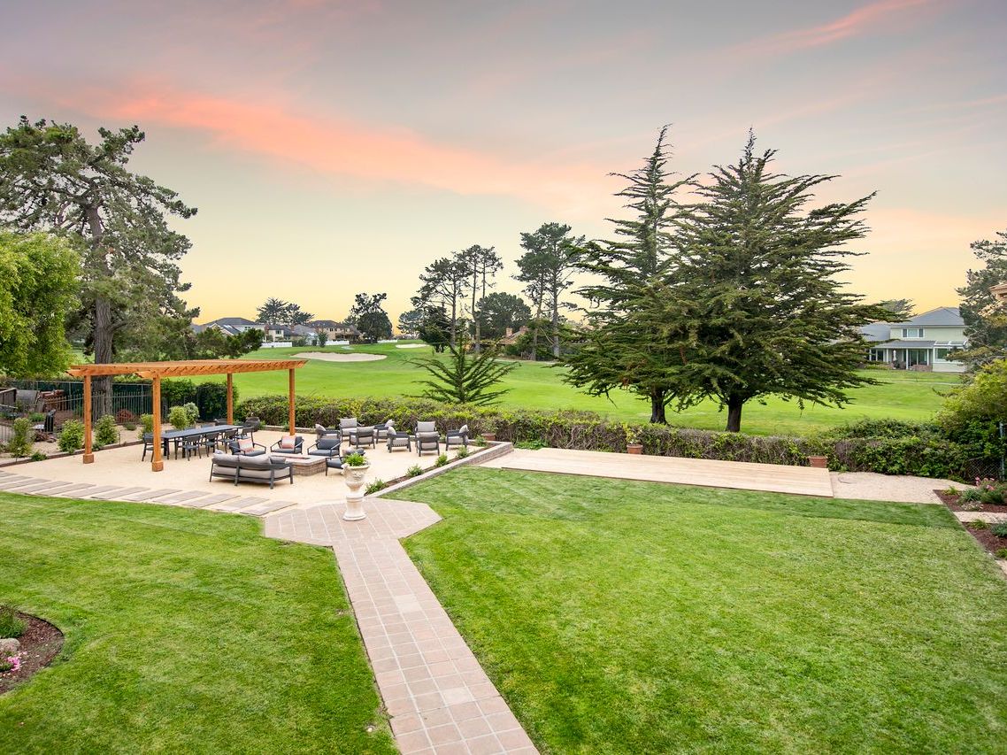 A landscaped backyard with a patio lounge, trimmed green lawn, flowering beds, tall trees, and a warm sunset sky.