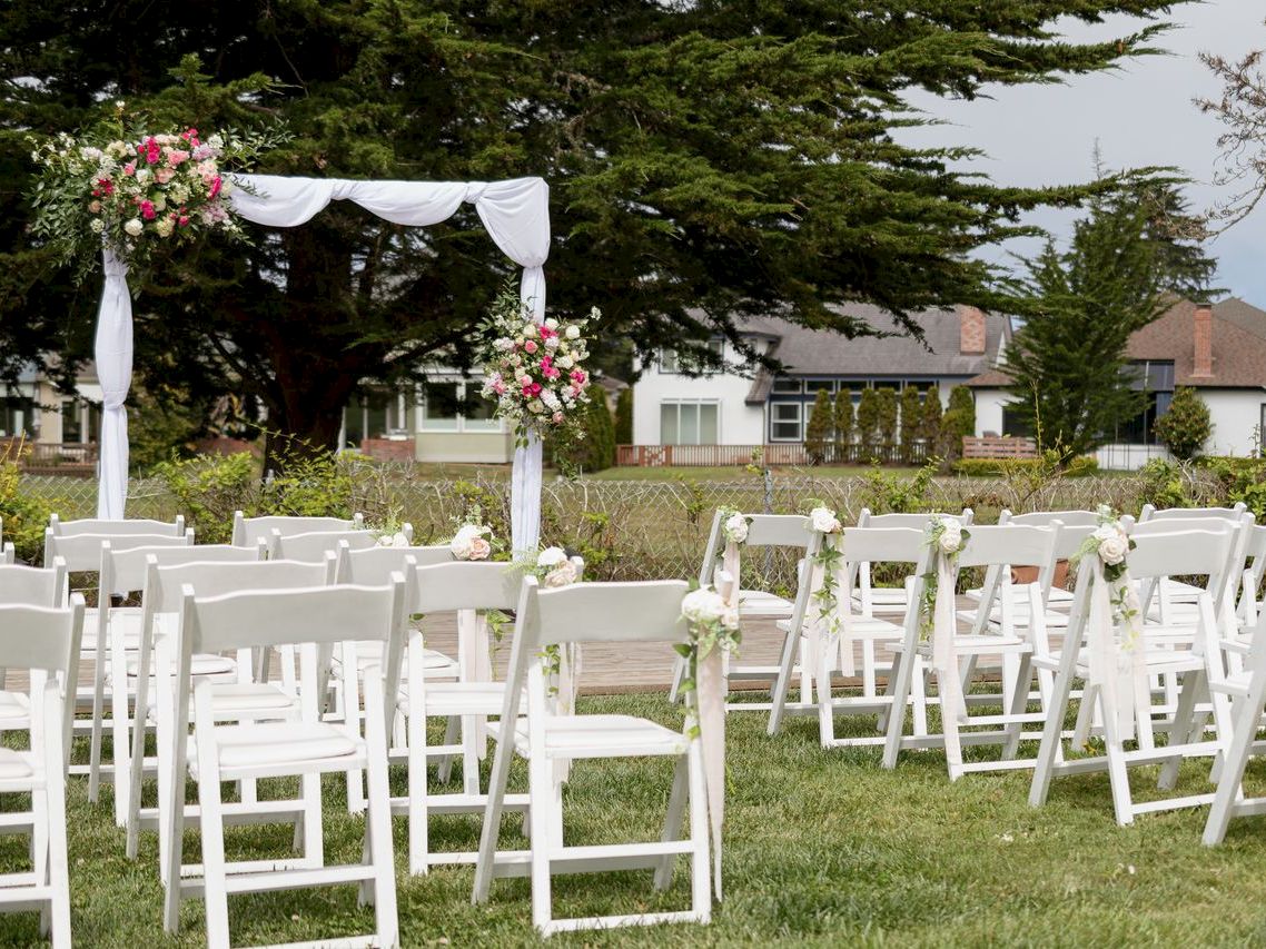 A wedding ceremony setup with white chairs on grass and a floral arch with pink and white flowers.