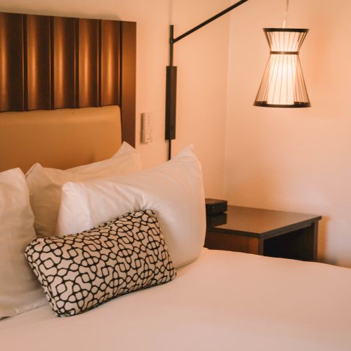A cozy bedroom corner with a bed, patterned pillows, a small wall lamp, and a wooden bedside table.