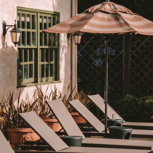 A cozy outdoor patio at night with a striped umbrella, lounge chairs, and a doorway wall lit by hanging lamps, above a garden hedge.