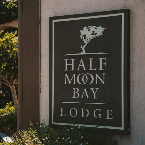 An outdoor plaque sign that reads &ldquo;HALF MOON BAY&rdquo; with an engraved tree emblem and &ldquo;LODGE&rdquo; below.