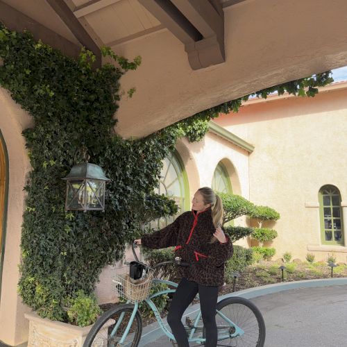 A woman in a dark jacket walks a turquoise bike past a stucco building covered in ivy and arched windows.