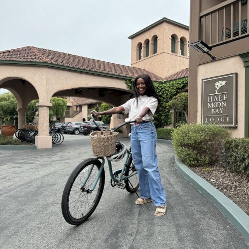 A smiling woman poses on a teal bicycle at the Half Moon Bay Lodge, with courtyard buildings and parked cars behind her.