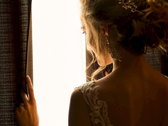 Backlit woman in lace dress stands by a curtain, peeking toward the light with one hand on the fabric.